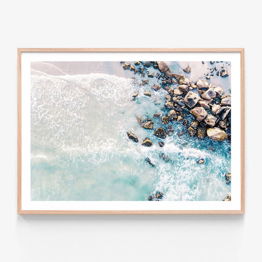 A framed artwork print of a beach scene with rocks and waves in blue tones.