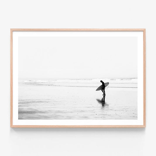 A framed print of a black and white photo of a surfer walking along the beach.