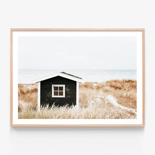 A framed photo wall art print featuring a small house on a coastal beach with tall grasses in the foreground, predominantly in black, white, and beige colors.