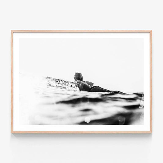 A framed art print featuring a black and white image of a person lying on a surfboard, paddling in the ocean.