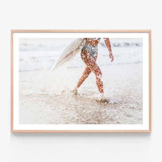 A framed photo wall art print featuring a woman carrying a surfboard while walking on a sandy beach with water splashing around.
