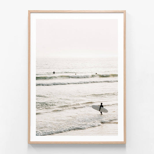 A framed photo wall art print featuring a coastal scene with a person holding a surfboard, standing in the shallow ocean waters with waves and a clear sky in the background.