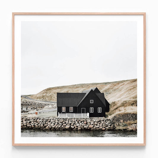 A framed photography print of a black wooden house with a thatched roof situated by a body of water, with a rocky shore in the foreground.