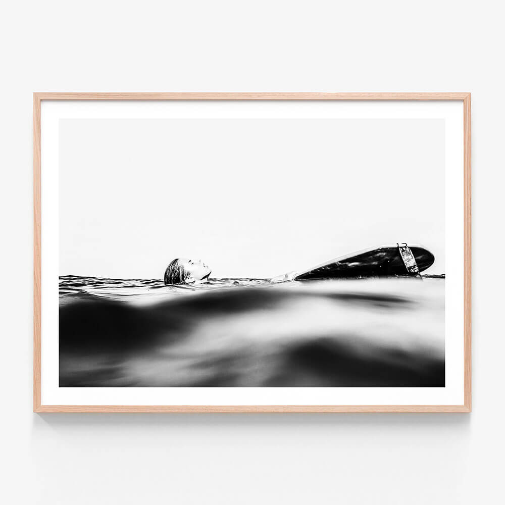 This framed art print captures the serene yet powerful essence of the ocean's coast. It features a black and white photograph of a surfboard partially submerged in the gentle waves, with the focus shifted slightly to the left.