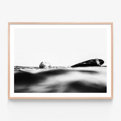 This framed art print captures the serene yet powerful essence of the ocean's coast. It features a black and white photograph of a surfboard partially submerged in the gentle waves, with the focus shifted slightly to the left.