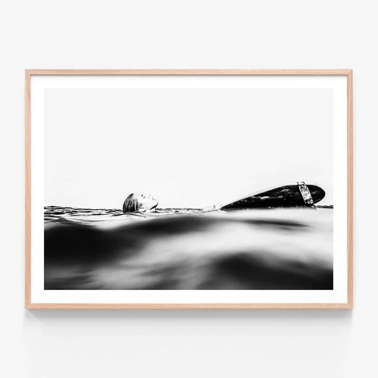 This framed art print captures the serene yet powerful essence of the ocean's coast. It features a black and white photograph of a surfboard partially submerged in the gentle waves, with the focus shifted slightly to the left.