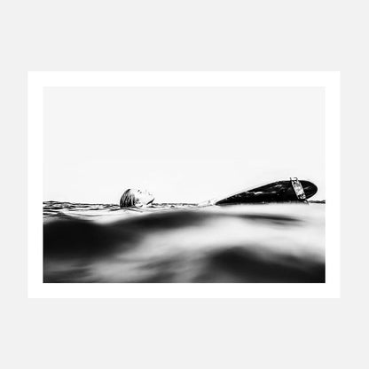 This black and white photography print captures the serene yet powerful essence of the ocean's coast. It features a surfboard partially submerged in the gentle waves, with the focus shifted slightly to the left.