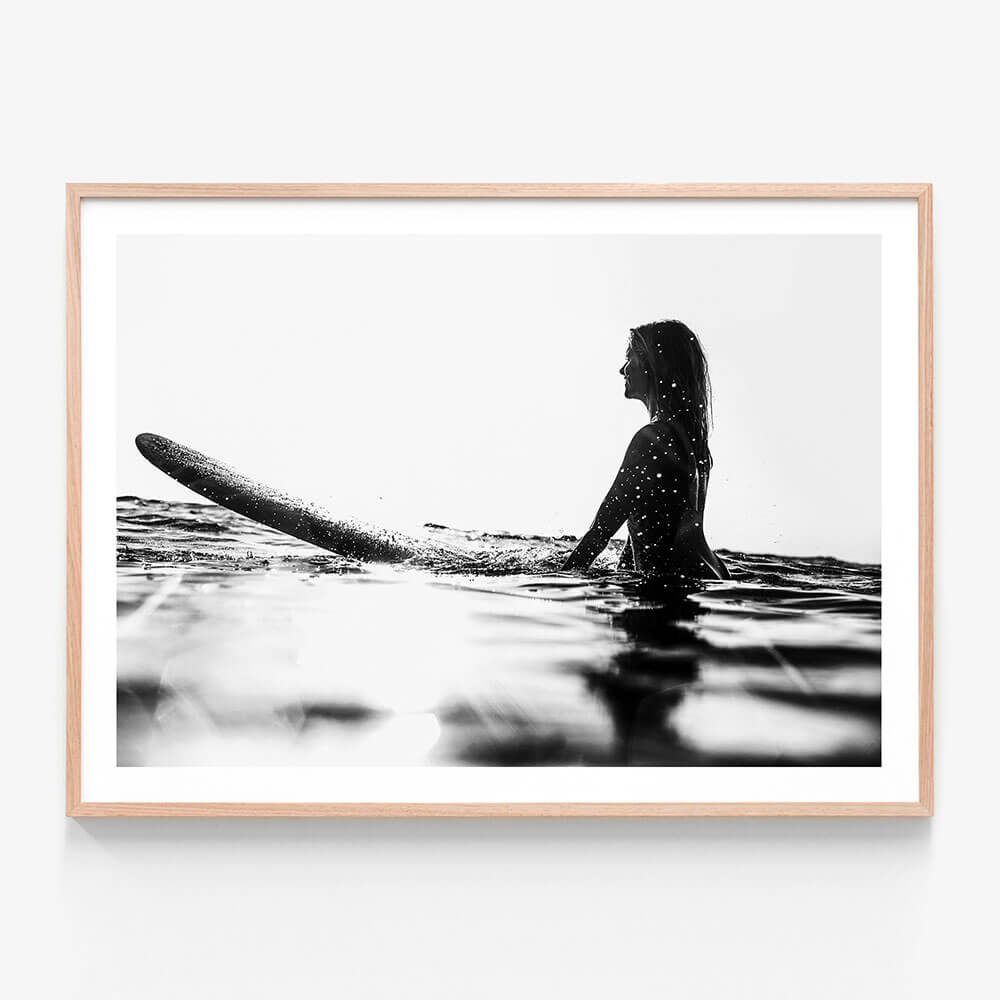A framed black and white wall art print photo of a person sitting on a surfboard in the ocean, looking off to the distance.