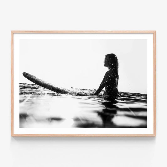 A framed black and white wall art print photo of a person sitting on a surfboard in the ocean, looking off to the distance.