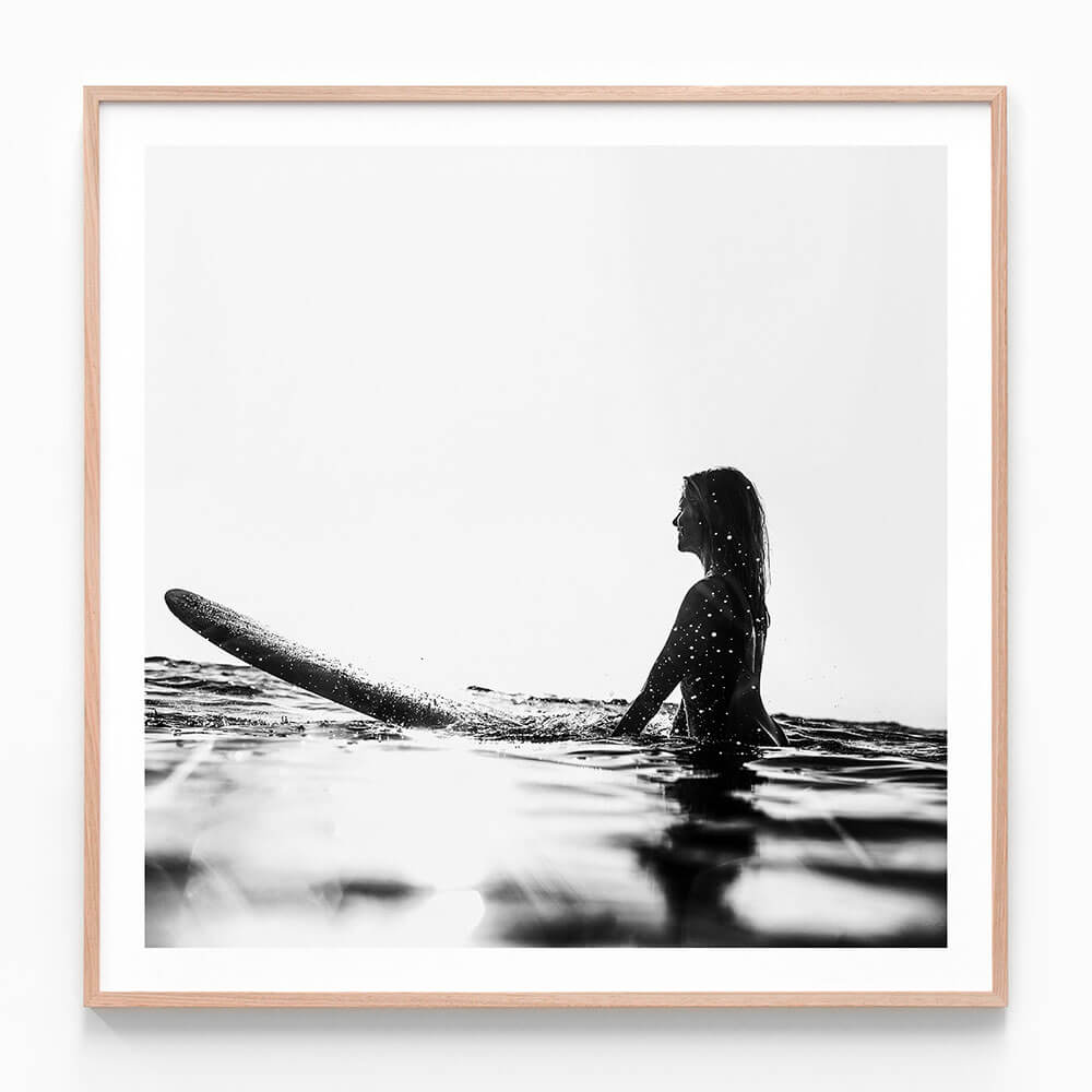 A framed print of a black and white photograph of a woman sitting on a surfboard in the ocean, with waves in the background.