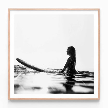 A framed print of a black and white photograph of a woman sitting on a surfboard in the ocean, with waves in the background.
