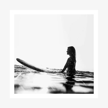 An art print of a black and white photograph of a woman sitting on a surfboard in the ocean, with waves in the background.