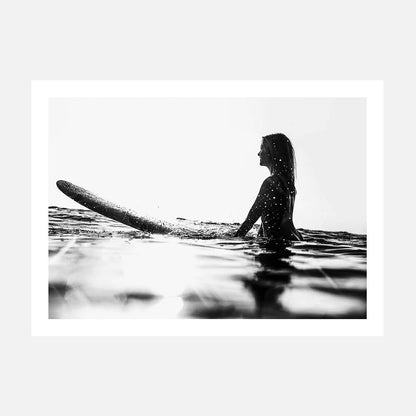 A black and white photo print of a person sitting on a surfboard in the ocean, looking off to the distance.