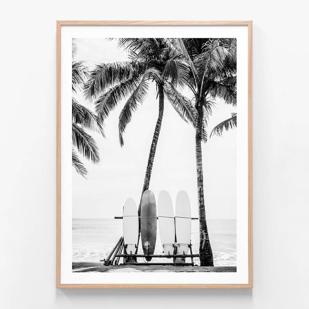 A framed black and white print of surfboards leaning against a lifeguard chair with palm trees in the background on a beach.