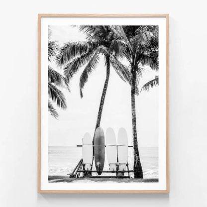 A framed black and white print of surfboards leaning against a lifeguard chair with palm trees in the background on a beach.