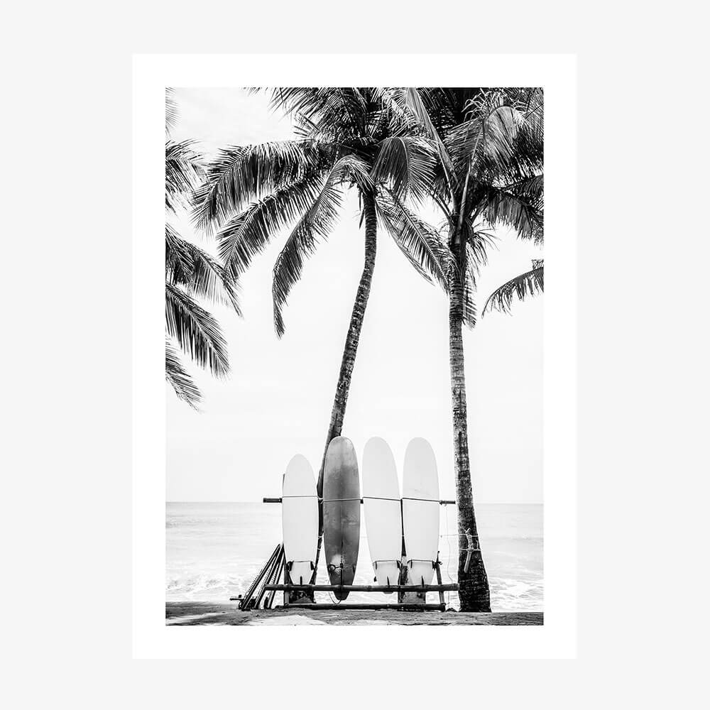A black and white photo art print of surfboards leaning against a lifeguard chair with palm trees in the background on a beach.