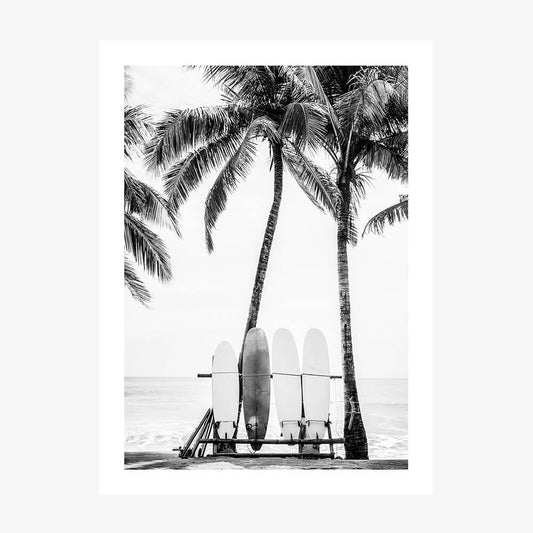A black and white photo art print of surfboards leaning against a lifeguard chair with palm trees in the background on a beach.