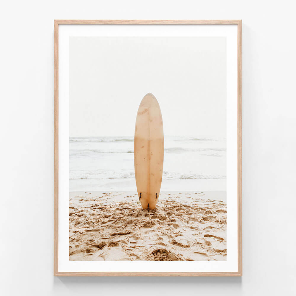 A coastal wall art framed print of a surfboard standing upright in the sand on a beach with the ocean in the background.