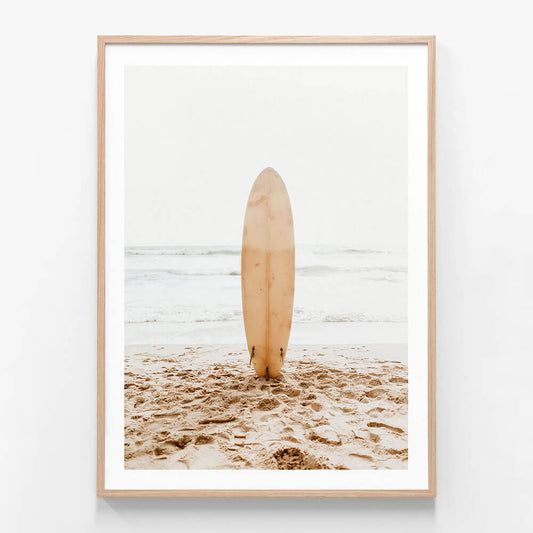 A coastal wall art framed print of a surfboard standing upright in the sand on a beach with the ocean in the background.