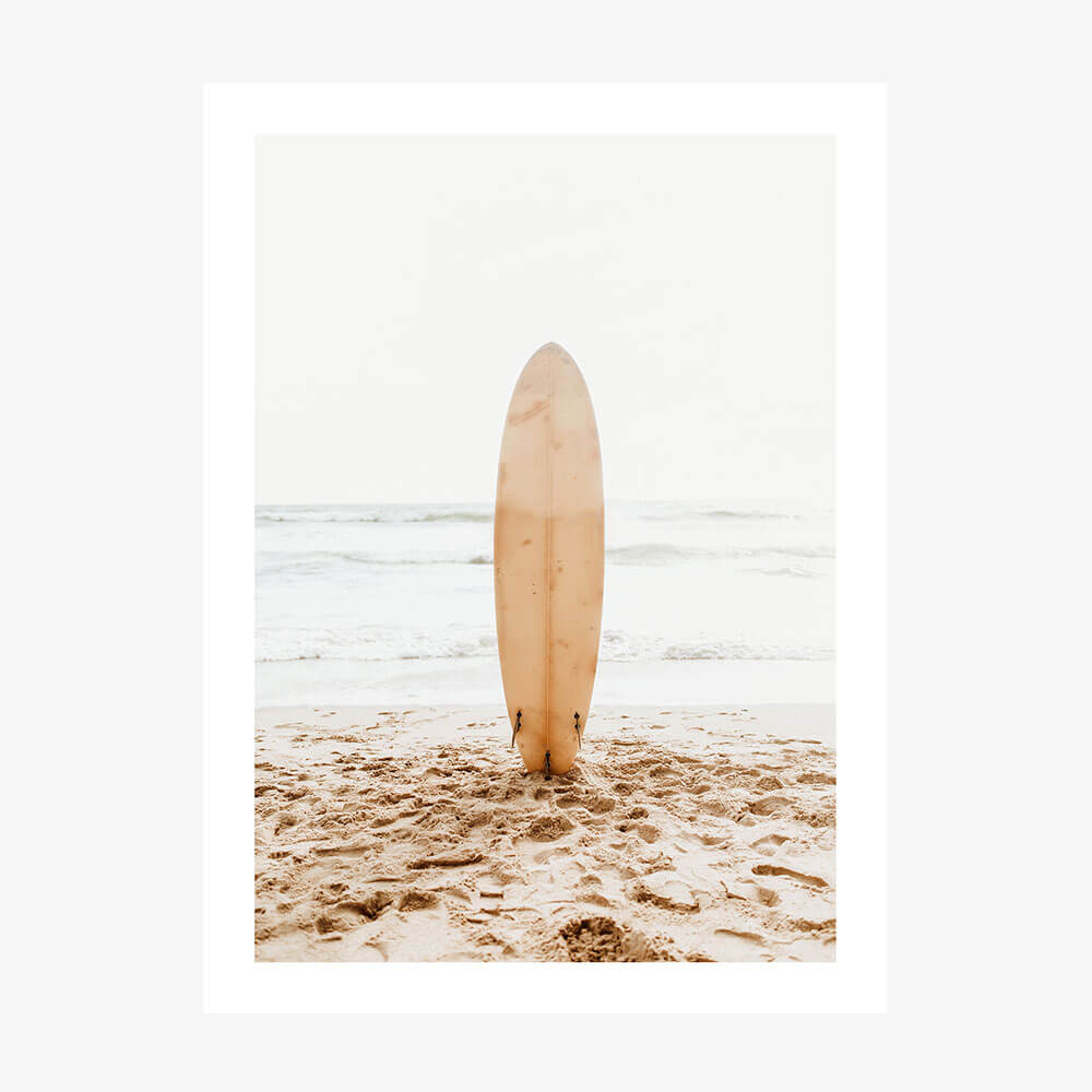 A photo wall art print of a surfboard standing upright in the sand on a beach with the ocean in the background.