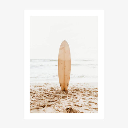 A photo wall art print of a surfboard standing upright in the sand on a beach with the ocean in the background.