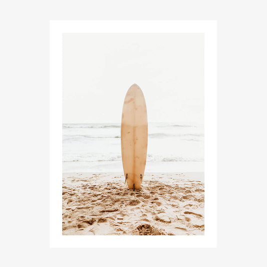 A photo wall art print of a surfboard standing upright in the sand on a beach with the ocean in the background.