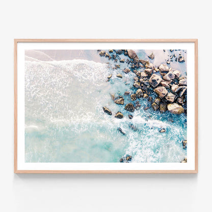 A framed artwork print of a beach scene with rocks and waves in blue tones.