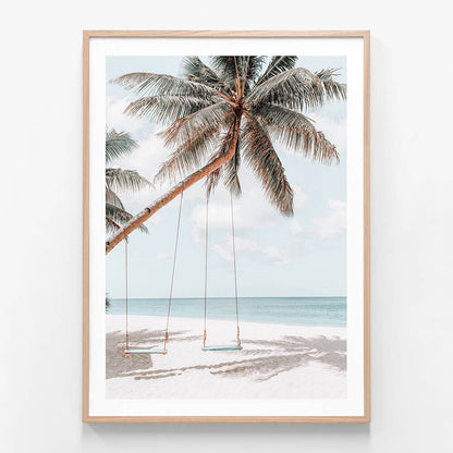 A framed wall art print featuring two swings hanging from a palm tree on a beach, with the ocean in the background.