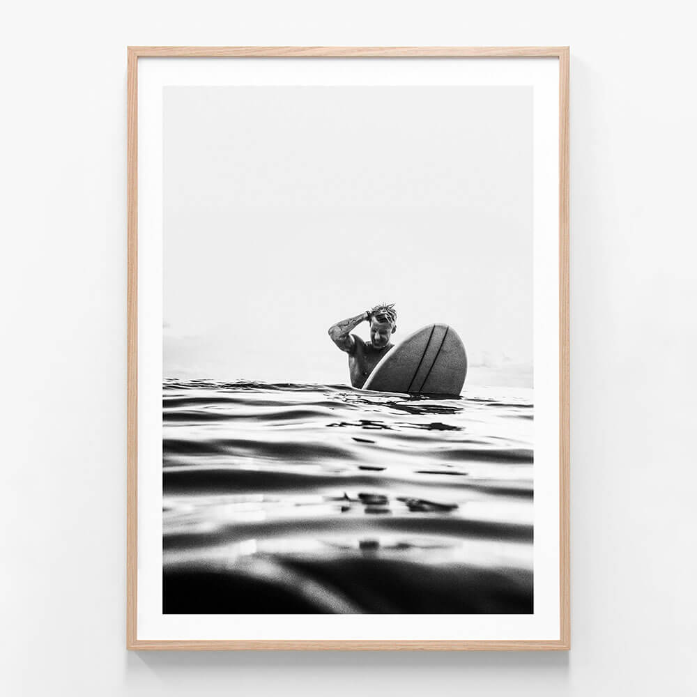 A framed art print featuring a black and white photograph of a surfer on a surfboard, with the surfboard predominantly in the frame against a backdrop of ocean waves.