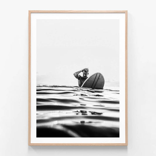 A framed art print featuring a black and white photograph of a surfer on a surfboard, with the surfboard predominantly in the frame against a backdrop of ocean waves.