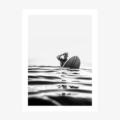 A black and white photograph print of a person on a surfboard, with the surfboard predominantly in the frame against a backdrop of ocean waves.