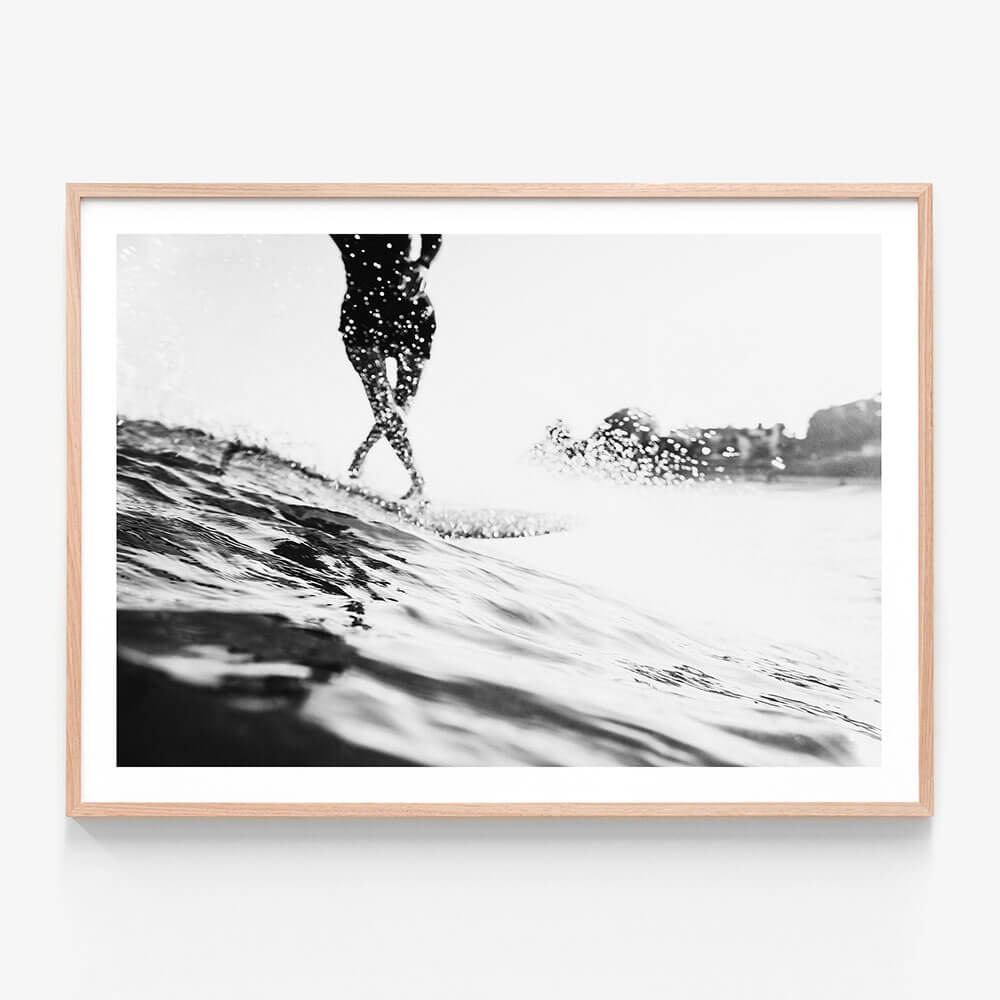 A framed black and white photo wall art print featuring a person surfing on a wave with a coastal background.