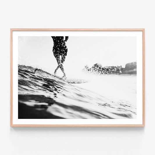 A framed black and white photo wall art print featuring a person surfing on a wave with a coastal background.