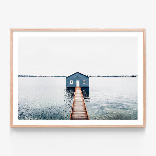 A framed wall art print of a small blue boat shed at the end of a pier over calm water, with a minimalist aesthetic.