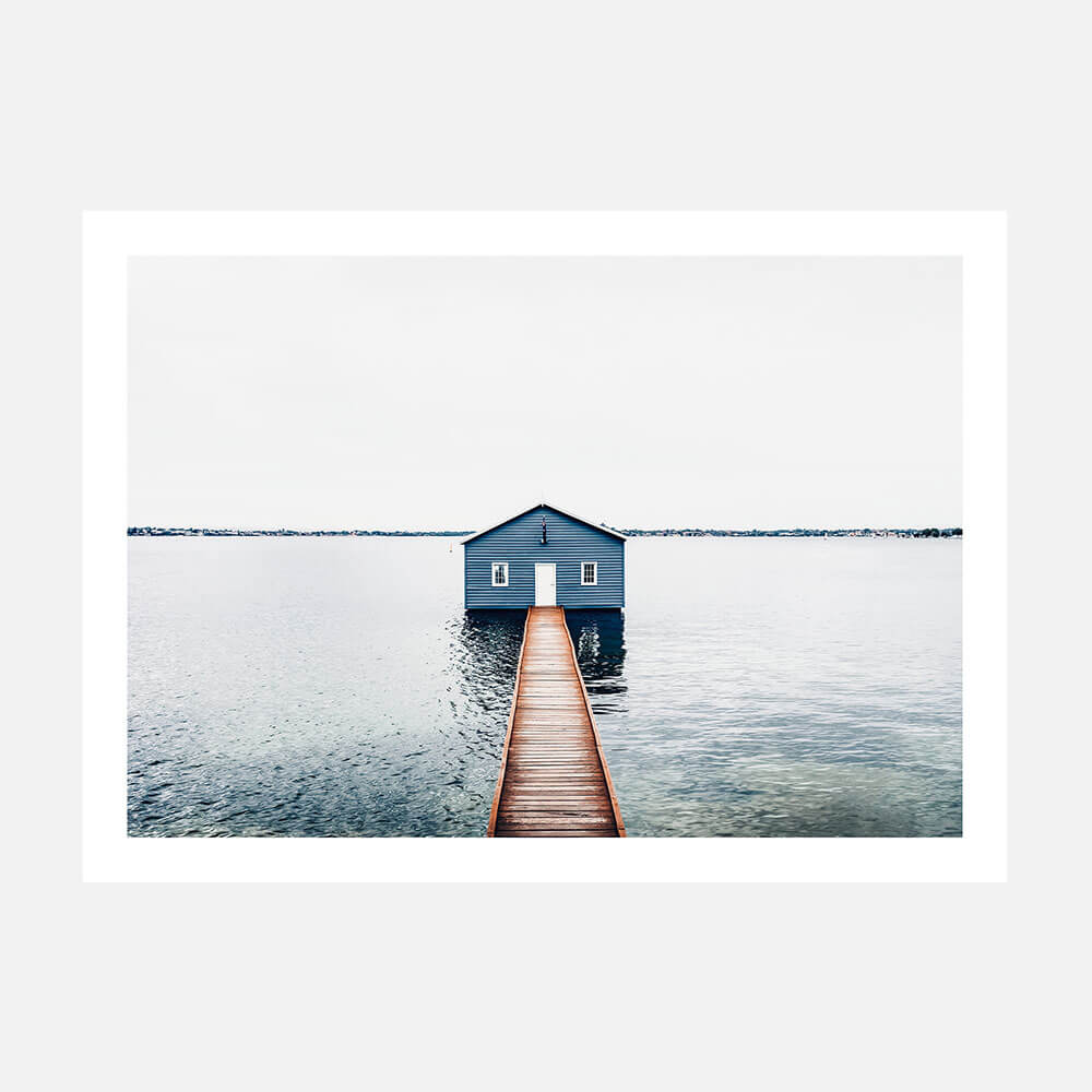 A photo wall art print featuring a small blue boat shed at the end of a pier over calm water, with a minimalist aesthetic.