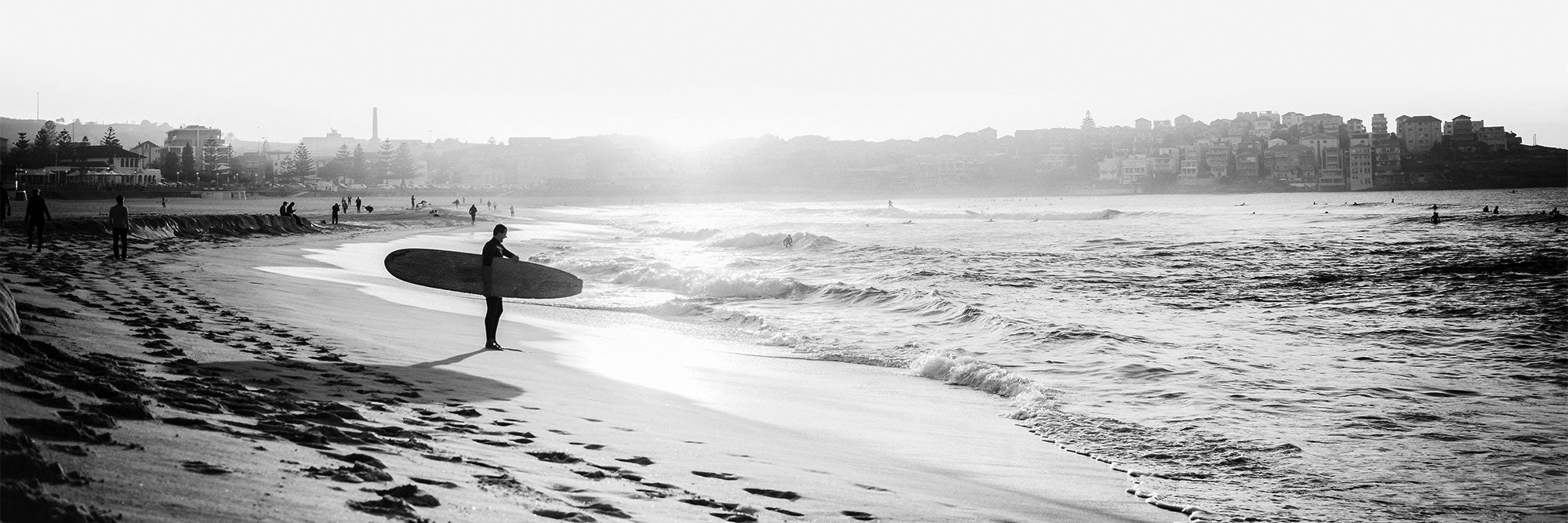 Black and white photographic wall art print of a person with a surfboard walking along Bondi Beach.