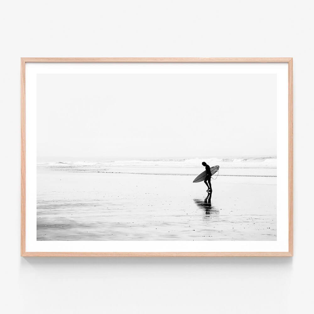 A framed print of a black and white photo of a surfer walking along the beach.