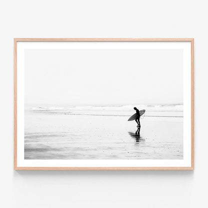 A framed print of a black and white photo of a surfer walking along the beach.