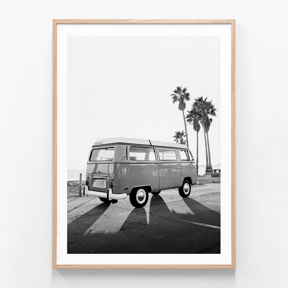 This black and white framed art print features a vintage photograph titled "Cali Van," showcasing a classic black and white image of a Volkswagen Type 2 van, commonly known as a "Kombi." The scene is set in a serene, sunlit environment with palm trees in the background, enhancing the iconic and idyllic vibe of the picture.