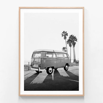 This black and white framed art print features a vintage photograph titled "Cali Van," showcasing a classic black and white image of a Volkswagen Type 2 van, commonly known as a "Kombi." The scene is set in a serene, sunlit environment with palm trees in the background, enhancing the iconic and idyllic vibe of the picture.