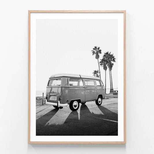 This black and white framed art print features a vintage photograph titled "Cali Van," showcasing a classic black and white image of a Volkswagen Type 2 van, commonly known as a "Kombi." The scene is set in a serene, sunlit environment with palm trees in the background, enhancing the iconic and idyllic vibe of the picture.