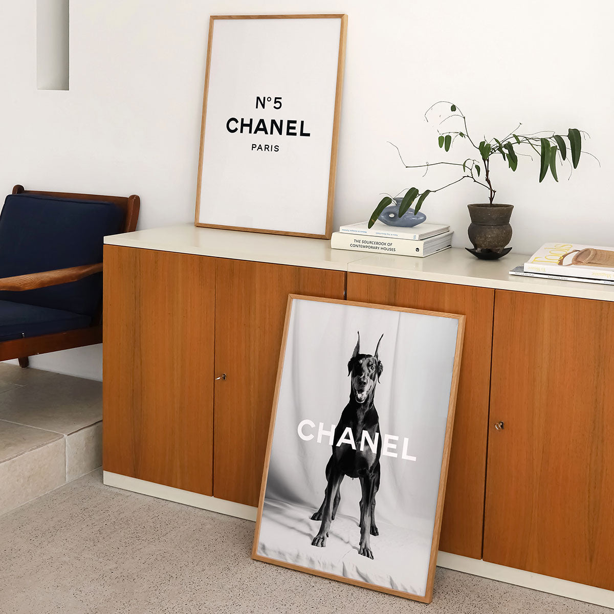 Framed Chanel poster with a Doberman print on a floor in a room with a wooden cabinet and plant.