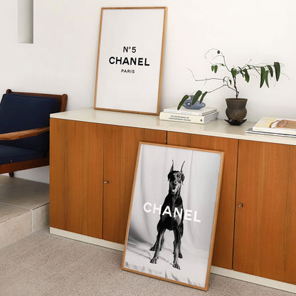 Framed Chanel poster with a Doberman print on a floor in a room with a wooden cabinet and plant.