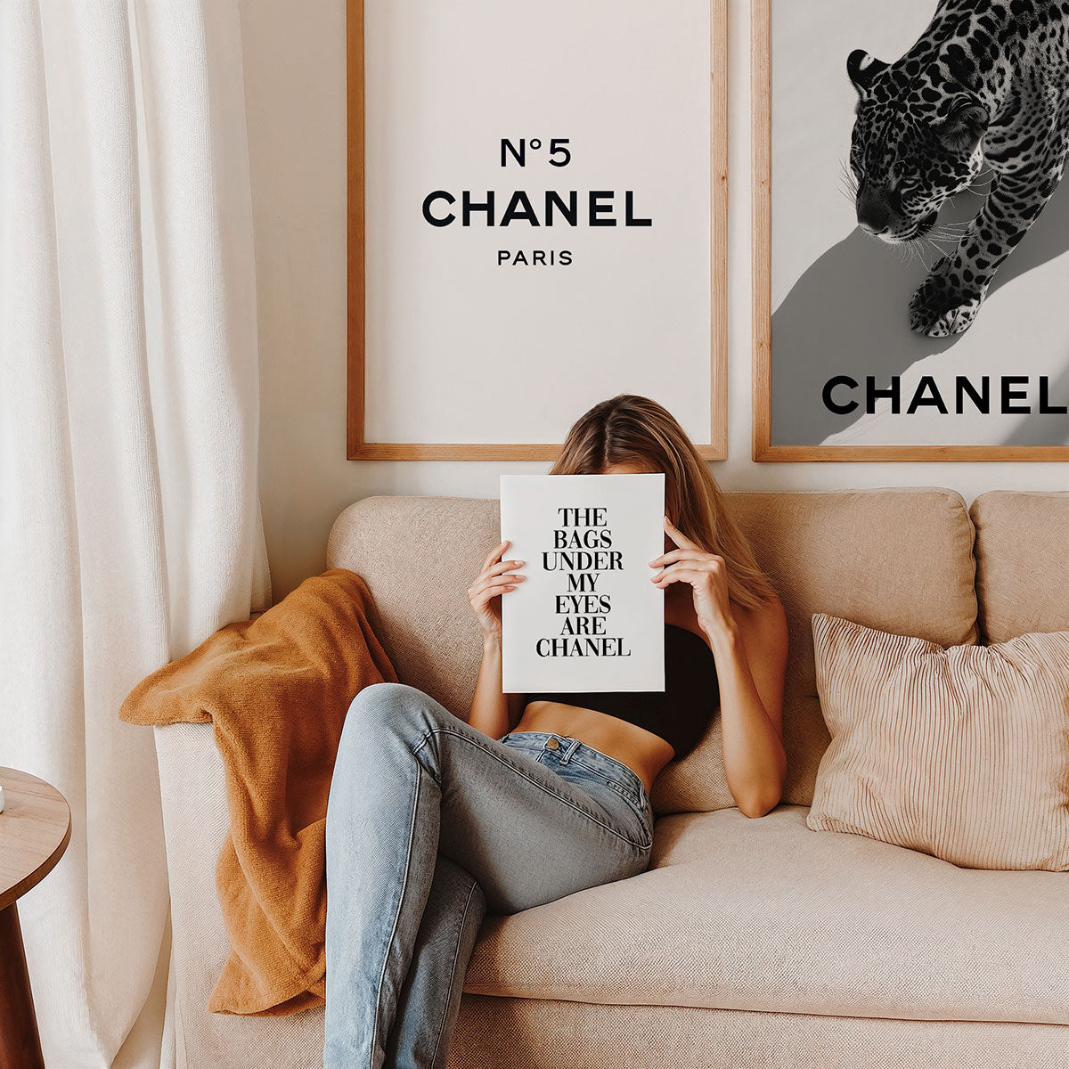 Woman sitting on a couch holding a Chanel poster, with fashion framed prints on the wall in the background.