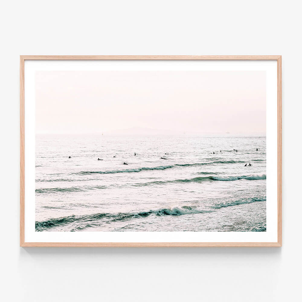 A oak framed print with a coastal surfboard theme, featuring a body of water with gentle waves and individuals on surfboards.