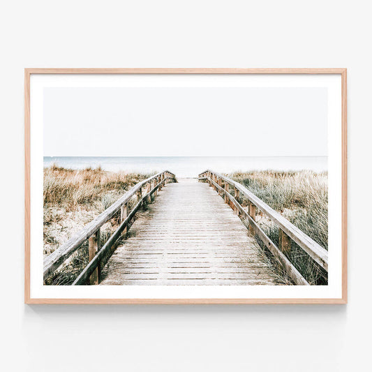 A print of a coastal boardwalk leading towards the ocean, with a bright sky and grassy dunes on either side.