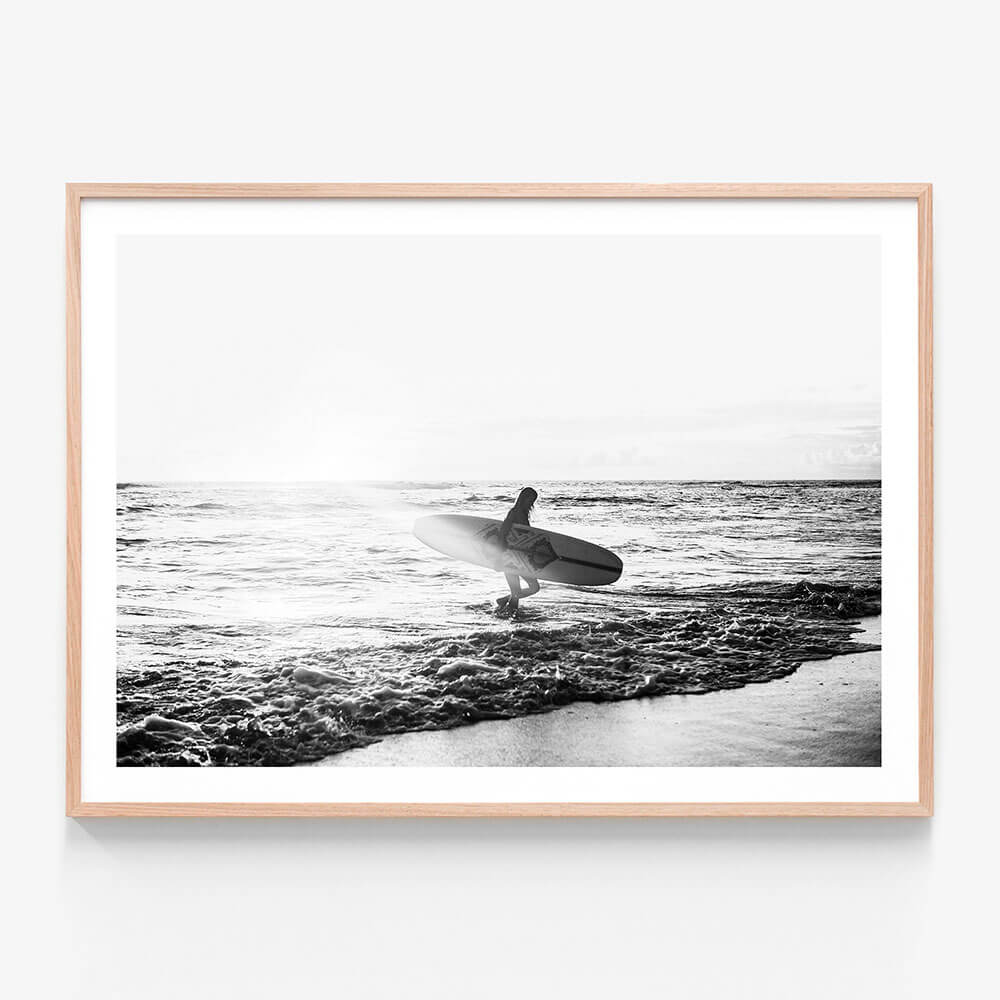 A framed print of a black and white photograph of a woman standing in the ocean holding a surfboard, with waves in the background.