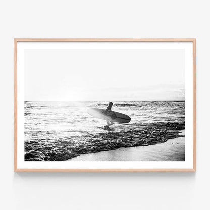 A framed print of a black and white photograph of a woman standing in the ocean holding a surfboard, with waves in the background.