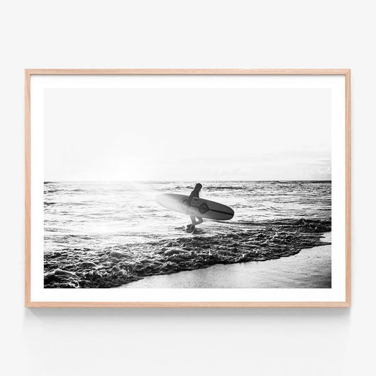 A framed print of a black and white photograph of a woman standing in the ocean holding a surfboard, with waves in the background.