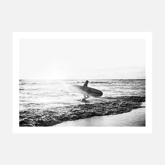 A black and white photography print of a person standing in the ocean holding a surfboard, with waves in the background.